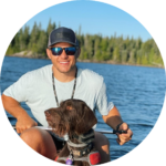 On Hook Agency's website: Justin Puetz, wearing sunglasses and a baseball cap, smiles in a boat with his brown dog, enjoying the tranquility of a tree-lined lake under a clear blue sky.