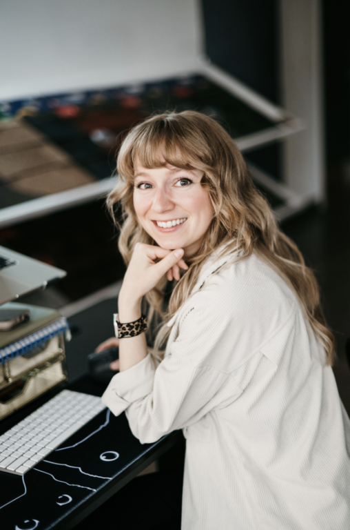 A woman with long, wavy hair sits confidently at her desk, ready to begin her work. Dressed in a light-colored shirt and watch, she glances over her shoulder with a smile. A laptop and vibrant board are visible in the background.