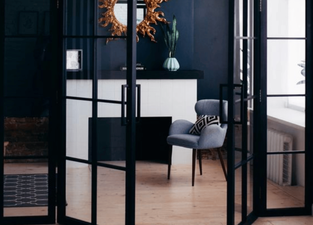 Transform your space with a modern living room design that captivates with its minimalist color palette. A striking dark blue wall complements a white fireplace, topped by an ornate gold mirror for added elegance. A mid-century armchair adorned with a geometric cushion sits on the wooden floor, all beautifully illuminated through glass-paneled doors.