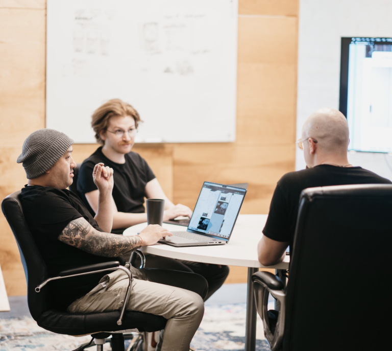 In a meeting room, three professionals collaborate on website design: one navigates the discussion using a laptop, another brings creative flair with tattoos and a beanie, while the third contributes insights from across the table. A whiteboard and large monitor enhance their strategic session.
