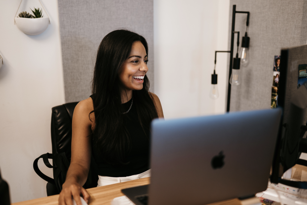 A woman with long dark hair in a black sleeveless top sits at a desk, deeply focused on managing Google Ads for home services. Her workspace is highlighted by gray acoustic panels, a modern lamp with exposed bulbs, and small potted plants.