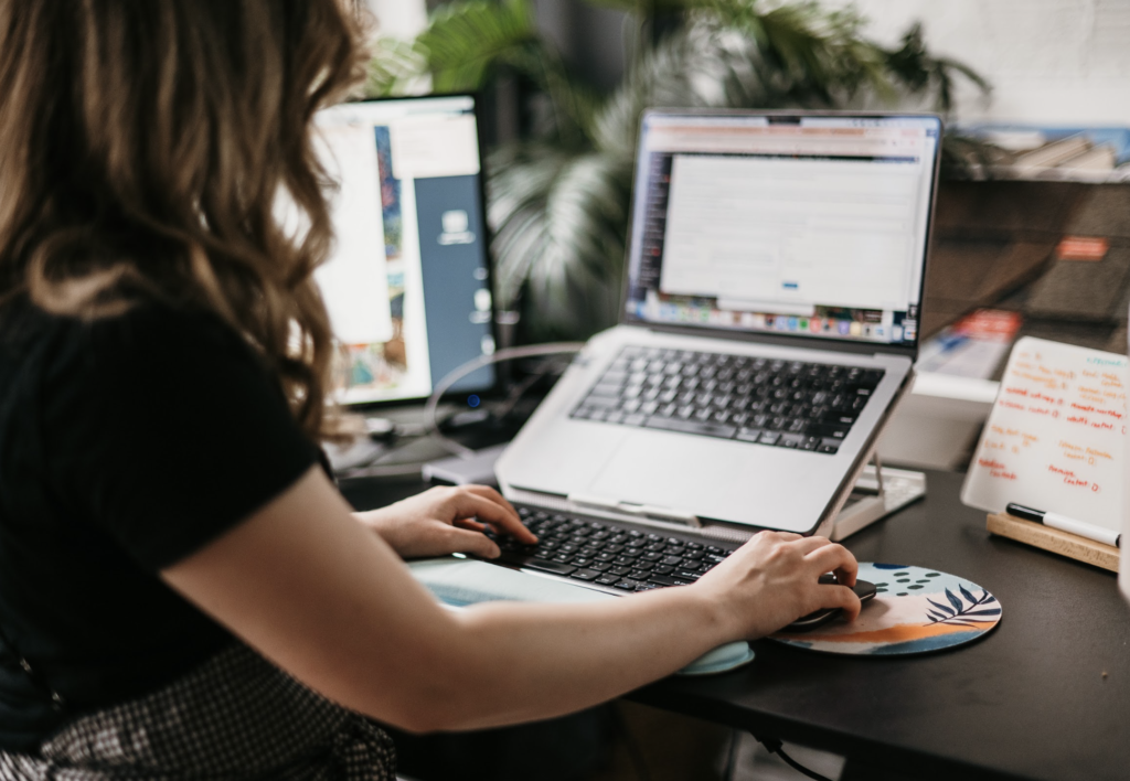 A woman with long hair concentrates intently on her desk, researching optimal blog post lengths for SEO. She types steadily on her laptop's keyboard, her hand resting on a brightly colored mouse pad amid office essentials and vibrant plants.