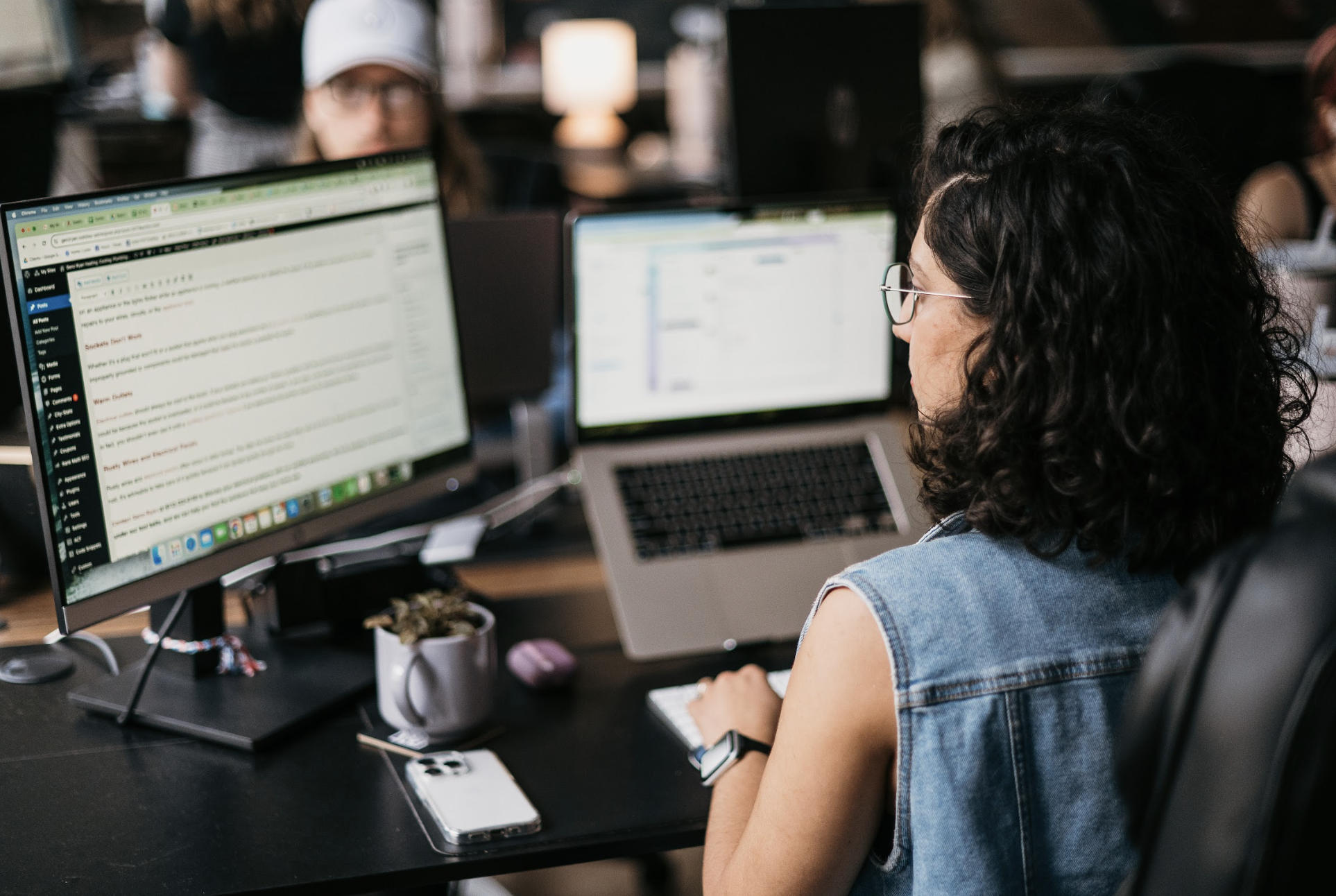 An expert in Home Services SEO, identifiable by their curly hair and glasses, efficiently optimizes content from a sleek laptop at their desk. The workspace features a large monitor displaying a text document, complemented by the charm of a smartphone and potted plant.