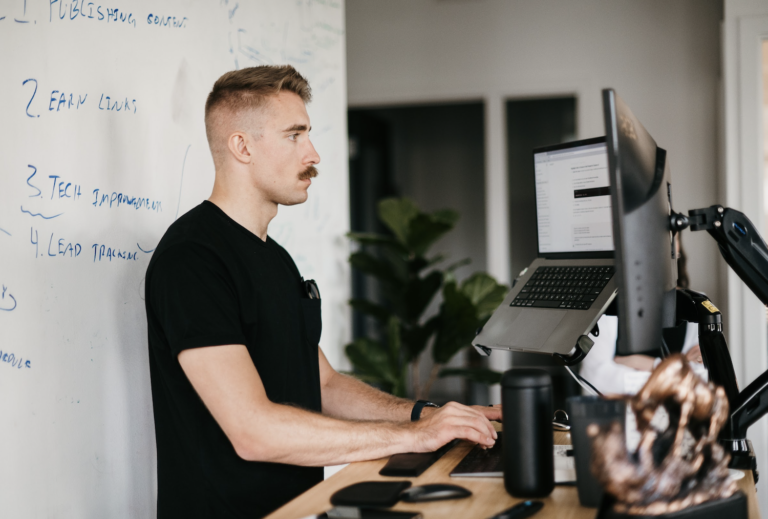 An individual with short hair and a mustache is diligently working at a standing desk. He types on a laptop attached to an elevated monitor, dressed in a black t-shirt. Behind him, a whiteboard displays detailed HVAC SEO strategies. A plant subtly adds to the workspace ambiance.