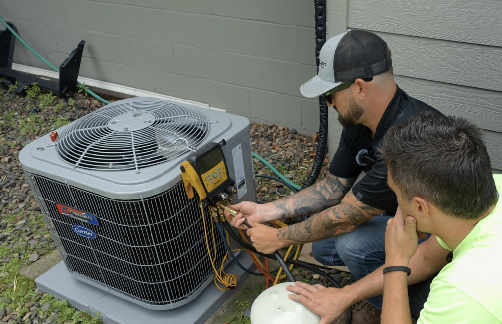 HVAC SEO - Two men are inspecting and working on an HVAC unit outdoors. One, in a cap and sunglasses, diligently adjusts the system while the other observes closely. Surrounded by tools and rocky ground next to a building, they're focused on optimizing local efficiency. Tim Brown on the right, runs Hook Agency helping clients with HVAC Local SEO, and Nick Thelen of Thelen Mechanical on the right shows him more about his business so Tim and his team can get better about at marketing that company efficiently and effectively. This strategy sets Hook Agency apart from it's many competitors because they get down in the trenches, and actually get stuff done with context from their clients industries, and is very niched allowing them to get better with every client. SEO HVAC Directories
