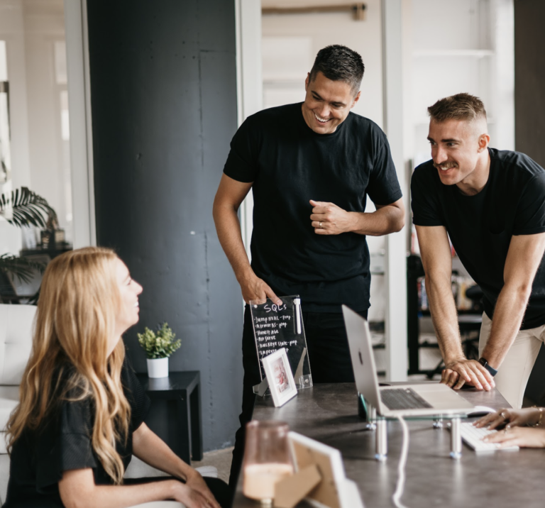 In a modern office filled with natural light, three professionals engage in a focused discussion on website design. Seated is a woman collaborating effectively with two standing men. Together, they exchange ideas around a desk equipped with a laptop, plant, and relevant documents.