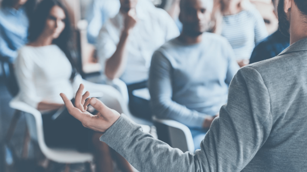 A speaker, seen from behind, gestures with a hand while addressing an attentive audience seated in chairs. With key principles at the forefront, the diverse audience is focused on enhancing professional visibility.