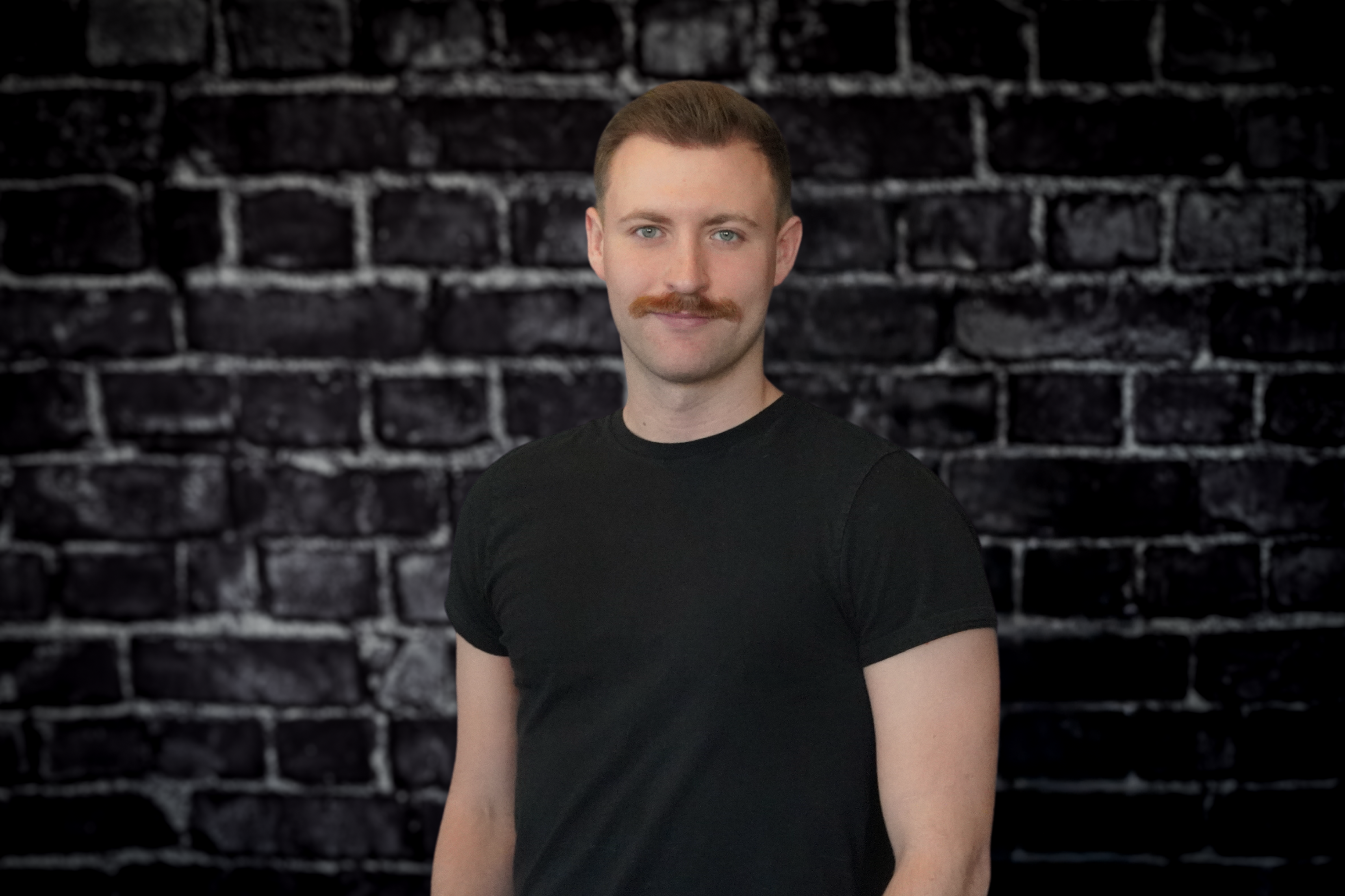 Jacob DeBoer, sporting a mustache and short hair, stands confidently against a dark brick wall. Wearing a black t-shirt, he gazes at the camera with a subtle smile.