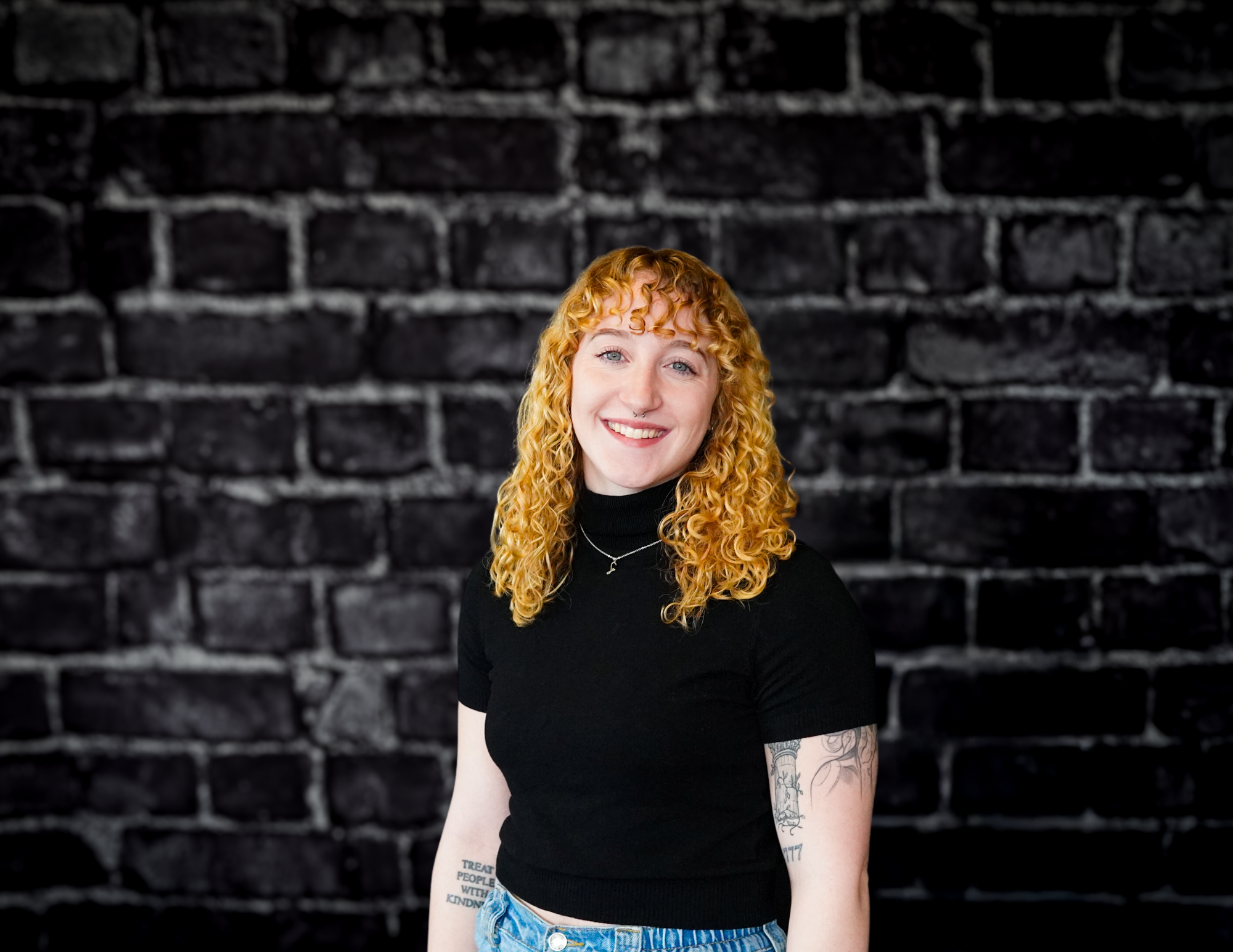 A professional with curly red hair, black turtleneck, and blue jeans smiles confidently before a dark brick wall on Hook Agency’s site.