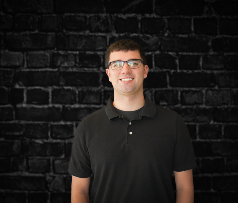 A man in glasses and a black polo shirt smiles confidently against a dark brick wall, echoing the minimalist style of artists such as Shameela Khan.
