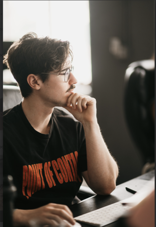 A man wearing glasses and a mustache sits thoughtfully at his desk, chin resting on hand. His black T-shirt boldly declares "POINT OF CONTACT" in red. The softly blurred office setting suggests a patio contractor project, with lighting accentuating his reflective demeanor.