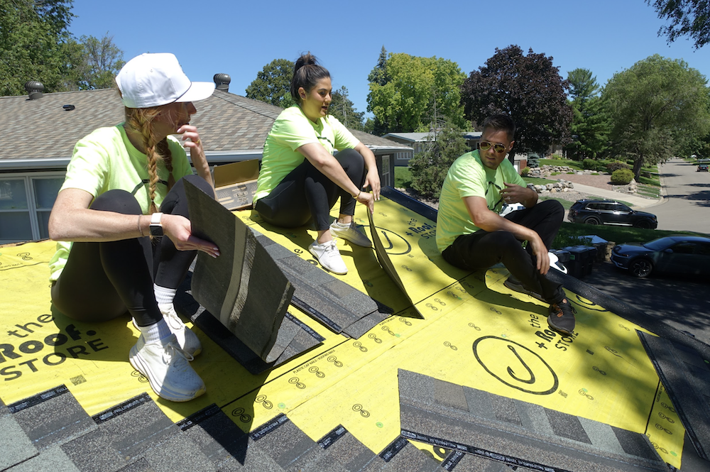 On a picturesque rooftop partially covered with yellow underlayment, three individuals in bright shirts discuss marketing strategies against a backdrop of trees and houses.