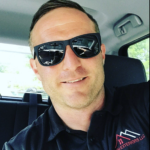Jacob Wadding, adorned in sunglasses and a black polo shirt featuring the "Exteriors LLC" logo with a roof design, is pictured inside a vehicle, smiling confidently at the camera. The car interior and a hint of the surrounding environment are visible behind him.