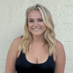 A smiling woman with blonde, wavy hair and a black sleeveless top stands confidently against a textured, light-colored wall.