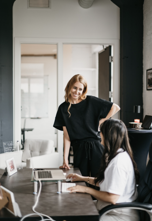 On Hook Agency's website, we feature a dynamic image of two women collaborating in a modern office setting. One woman stands, smiling and leaning slightly forward, while the other sits typing on her laptop. They are actively engaged in a productive discussion about website design strategies.