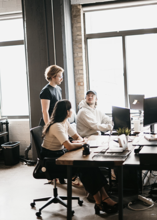 In a modern office setting, three professionals collaborate around computer desks. A man in a black shirt actively discusses SEO strategies with another seated man in a white hoodie and cap, while a woman nearby engages in the conversation. Large windows flood the space with natural light.