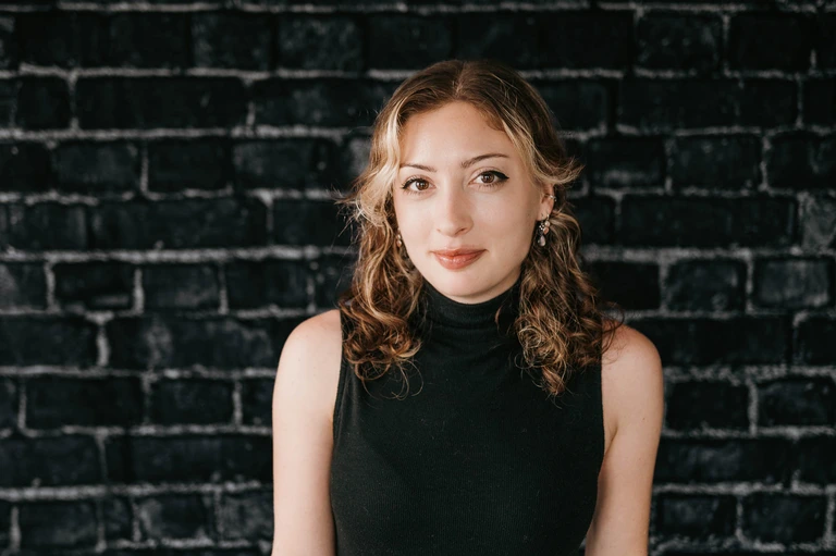 A woman with curly hair, dressed in a black sleeveless turtleneck, confidently stands against a dark brick wall. Emulating Tim Brown's assured presence, she looks directly into the camera with a subtle smile.