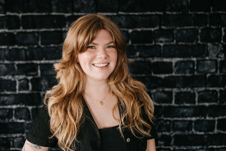 Tim Brown stands confidently in front of a textured black brick wall, exuding style and warmth. With long hair, a dark outfit, and a delicate necklace, their presence is undeniably captivating.