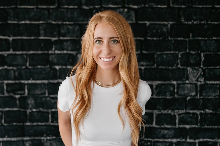 Tim Brown, with long reddish-blonde hair, stands confidently before a black brick wall, wearing a white T-shirt and a gold necklace.