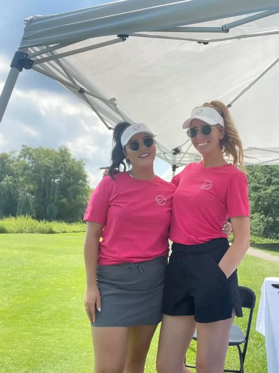 Two women, dressed in matching pink shirts, sunglasses, and white visors, smile confidently under a sunlit canopy on the golf course. Their demeanor suggests they are celebrating successful careers. Lush trees and a grassy expanse provide a picturesque backdrop.