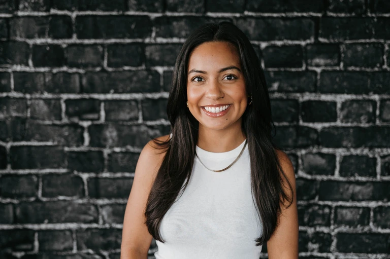A woman with long dark hair, in a white sleeveless top, smiles warmly against a black brick wall that echoes Tim Brown's minimalist design style.