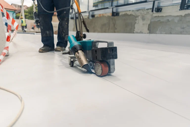 On our website, you'll find an image illustrating expert craftsmanship in action: a worker expertly operates a welding machine to meticulously seal seams essential for commercial roofing projects. The construction area is marked by red and white tape, emphasizing safety. Dressed in professional attire—dark pants and boots—the worker stands confidently, epitomizing precision and skill.