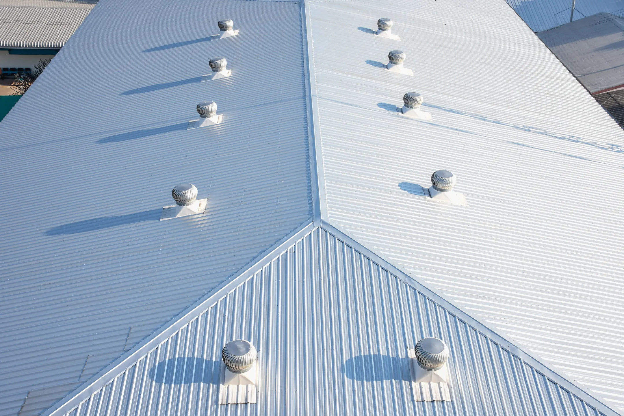 Capture the essence of industrial precision with this aerial view of a commercial rooftop. Featuring an expansive metal roof accented by evenly spaced round ventilation turbines along its ridges, the bright white surface casts striking shadows, highlighting the structure's symmetrical design.