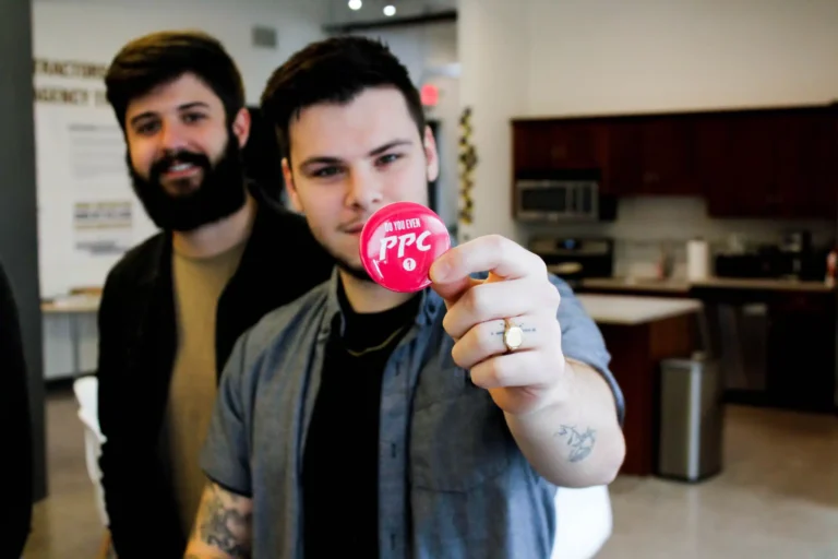 In an office-like setting, a tattooed individual confidently presents a red "PPC" button, symbolizing their readiness to address commercial roofing challenges. A second person stands slightly blurred in the background.