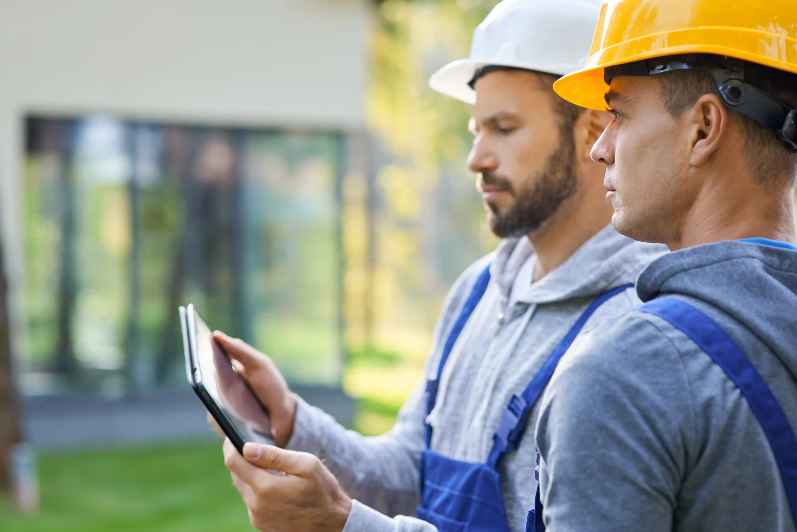 Two construction workers in helmets and blue overalls stand outside, intently reviewing critical marketing strategy data on a tablet. A construction site looms in the blurred background.
