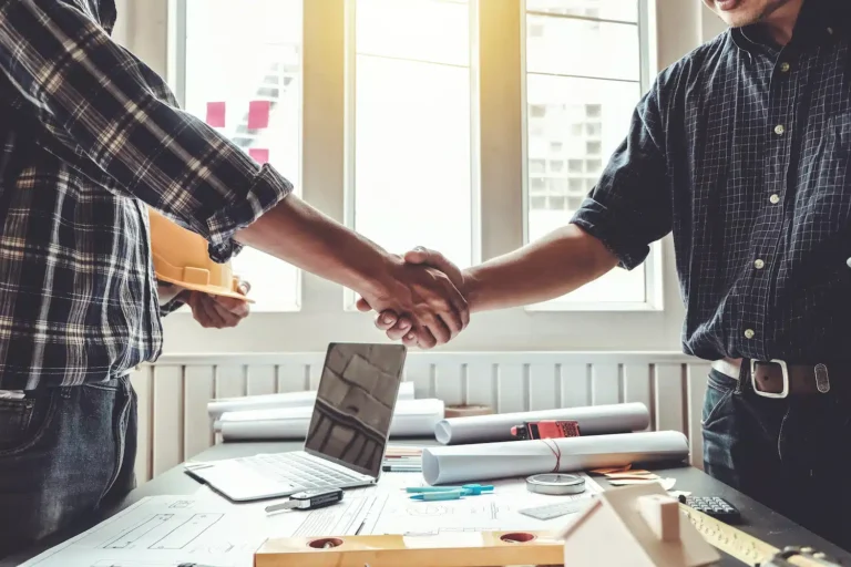 On a sunlit table filled with architectural plans, a laptop, and drafts, two professionals in checkered shirts shake hands, sealing a contractor collaboration.