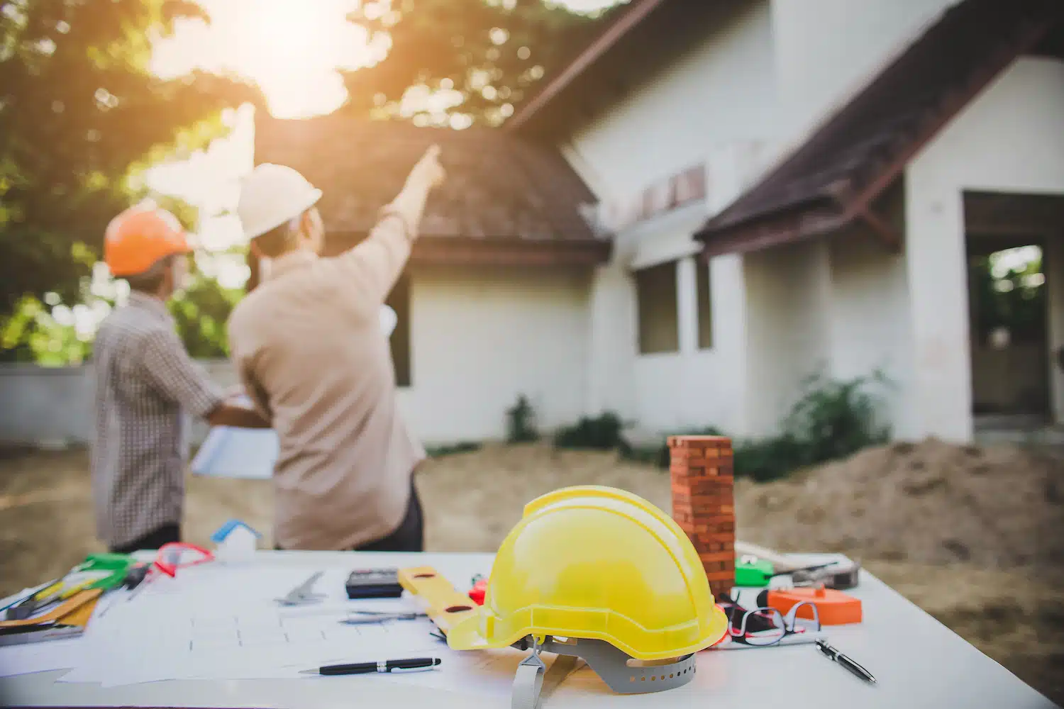 On the Hook Agency website, an image depicts two construction workers in hard hats standing outside a house. One points towards the building, illustrating active engagement in their work. In the foreground, a table displays essential tools of the trade: a yellow hard hat, blueprints, and other equipment—all representing contractor marketing under a bright sunlit sky.