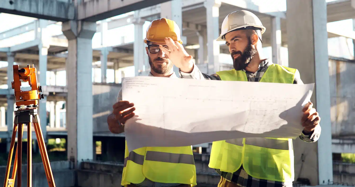 On a construction site, two workers in hard hats and reflective vests study blueprints, likely under a skilled contractor's guidance. One points out details as the other attentively observes. A surveying tool in the foreground captures vital site information.