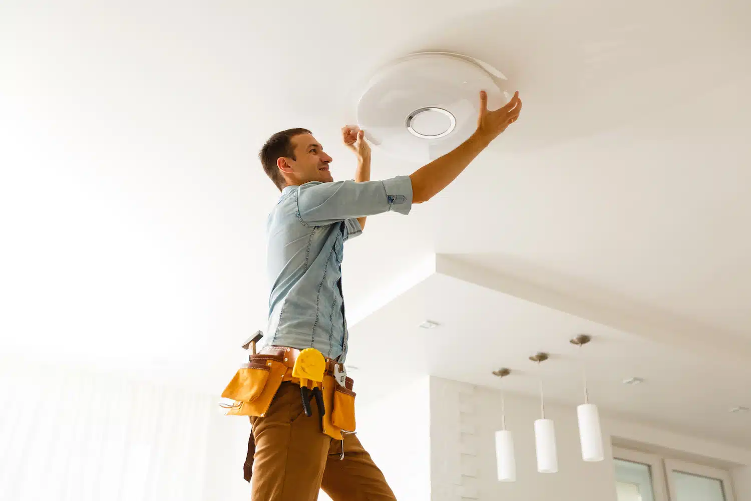A skilled contractor in a blue shirt and tool belt installs a round ceiling light fixture from a ladder, exemplifying precision. The room's sleek design—with bright white walls and modern hanging lights—sets the standard for chic website aesthetics.