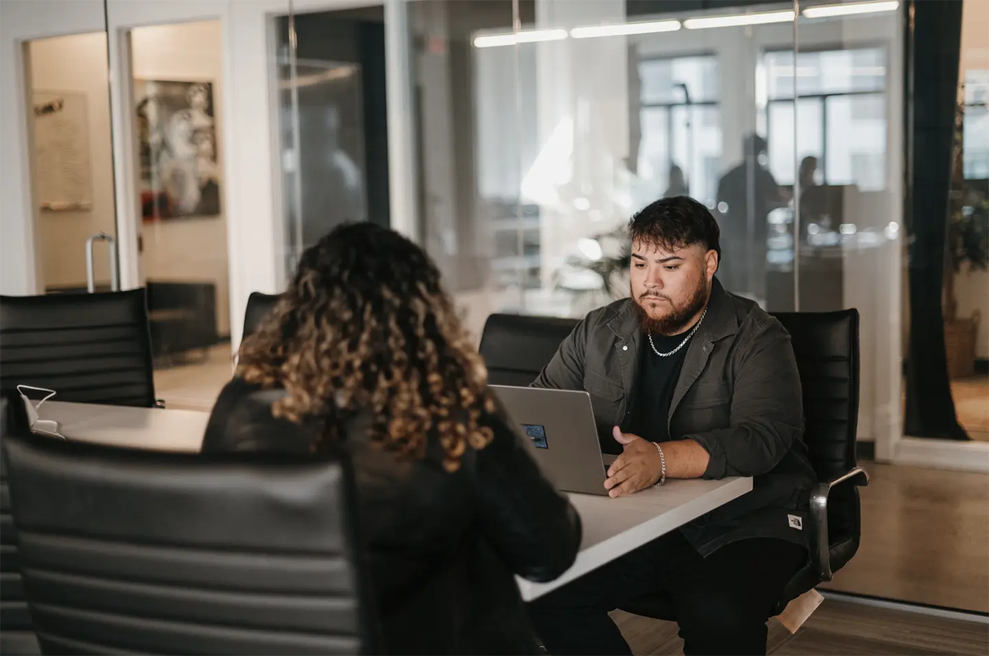 In a modern office surrounded by glass walls and professional seating, two individuals are focused on their tasks. One optimizes processes through a laptop, while the other listens attentively.