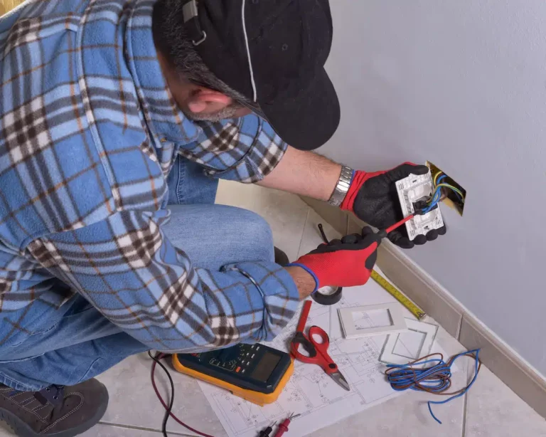 An expert electrician, equipped with a plaid shirt, cap, and red gloves, skillfully installs or repairs an electrical outlet. Surrounded by essential tools such as a multimeter and wiring diagrams, this professional exemplifies mastery in electrician PPC.