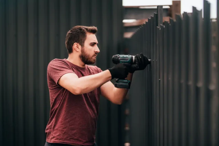 On Hook Agency's website, a professional in a red T-shirt and black gloves expertly uses a power drill to assemble vertical metal panels outdoors. This imagery exemplifies the precision and expertise expected from a leading fence company, with the dark fence extending into the background showcasing his unwavering focus.