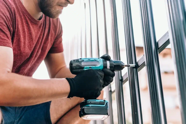A skilled craftsman, wearing a red T-shirt and black gloves, expertly secures metal bars with a cordless drill in an outdoor fence project. His precise technique underscores the quality and reliability of our trusted fence company.
