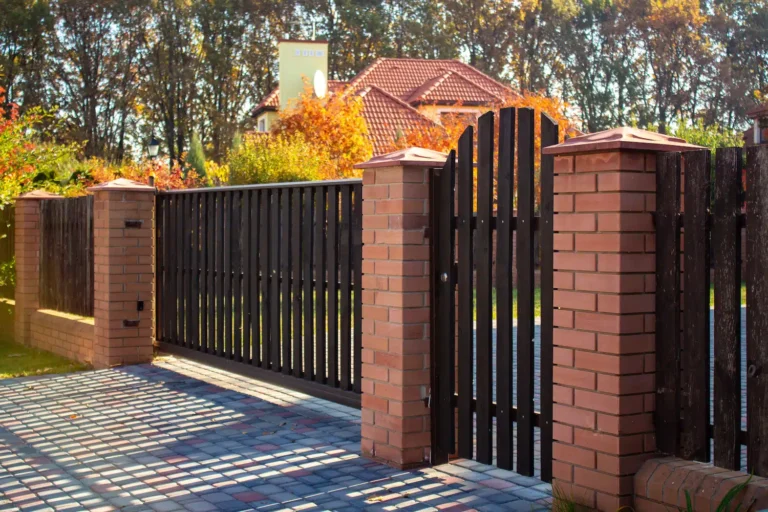 A black metal gate, surrounded by brick pillars, opens to a cobblestone driveway leading to a house with a red tiled roof. In the background, trees adorned in autumn foliage stand out against a clear blue sky, with sunlight casting gentle shadows on the path.