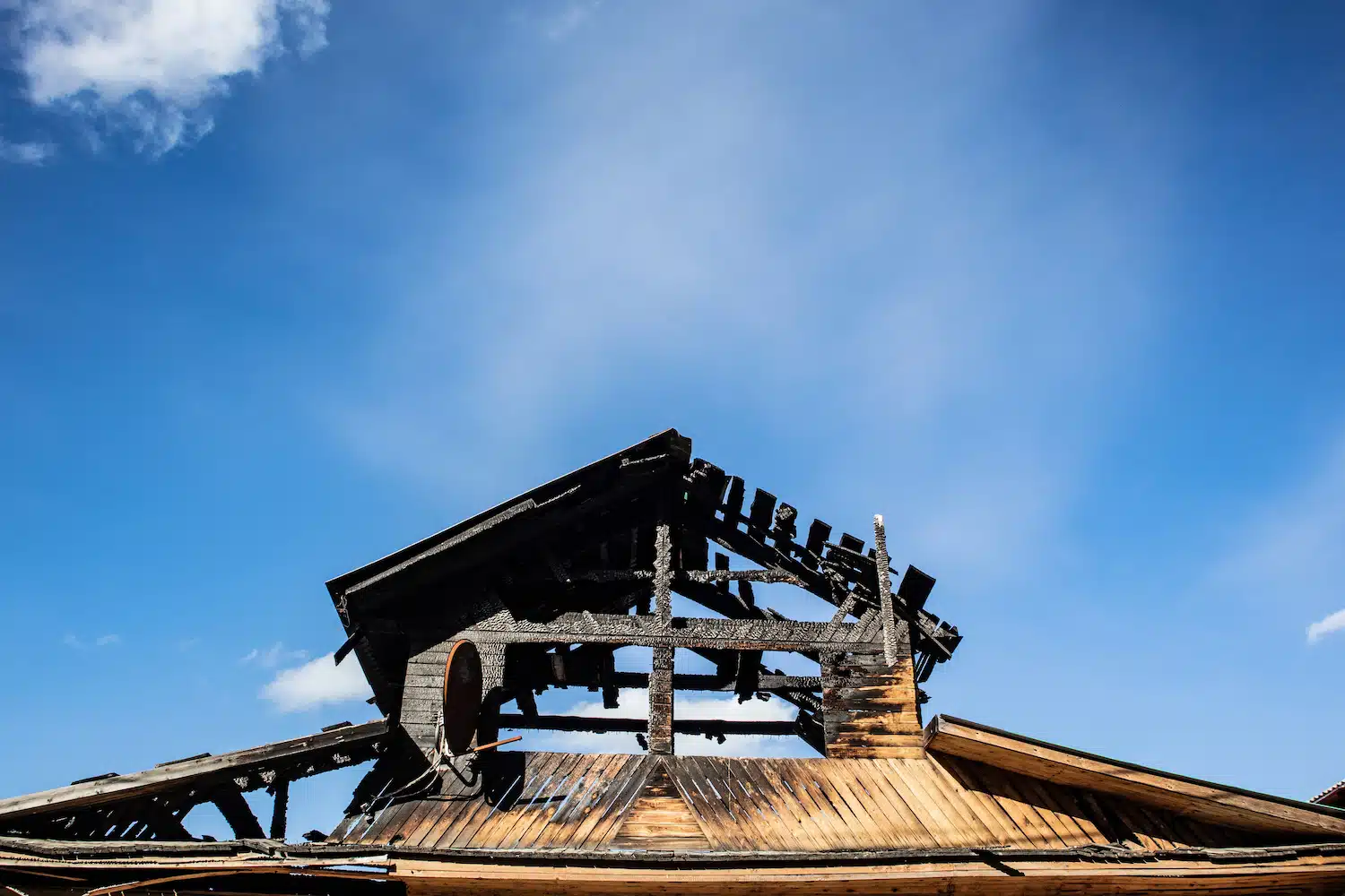 A fire-damaged wooden structure stands under a clear blue sky, awaiting restoration. The partially collapsed, charred roof exposes blackened beams, while drifting clouds suggest hope beyond the destruction.