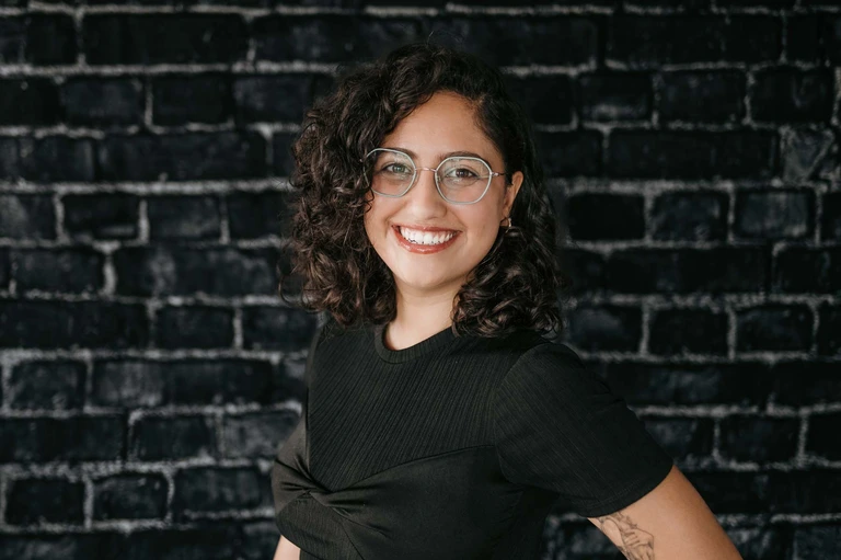 A smiling individual with curly hair and glasses, resembling Tim Brown, stands confidently in front of a textured dark brick wall, dressed in a black shirt.