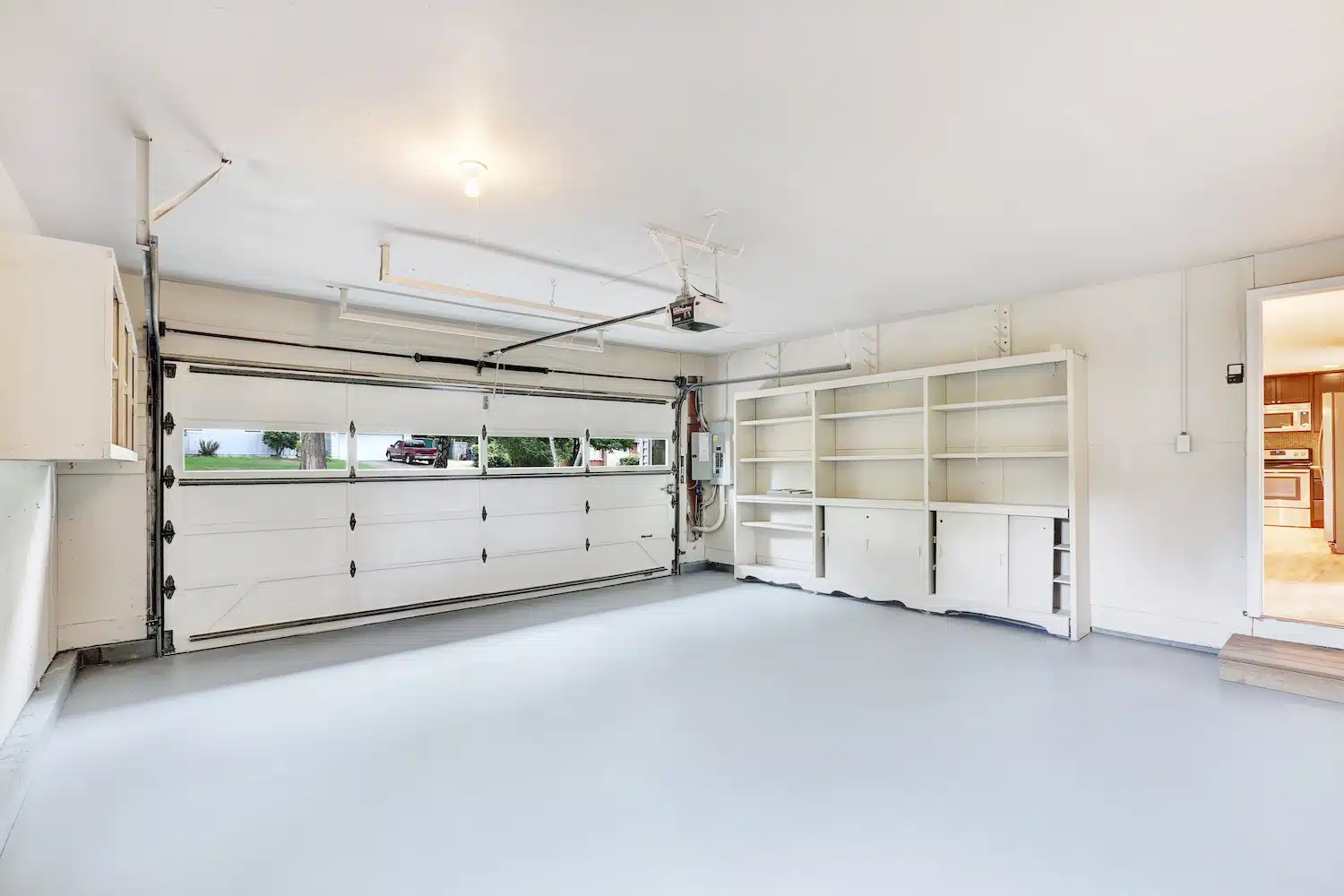 Explore our expertly transformed garage featuring a premium-coated, sleek polished concrete floor. The space is complemented by pristine white walls and ceiling, built-in side shelving units, and a closed double garage door adorned with top windows. A convenient doorway offers access to an adjoining room.