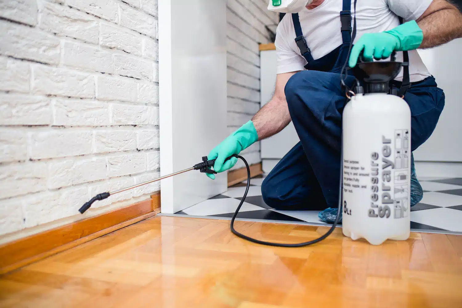 On Hook Agency's website: A professional in protective gear expertly applies pest control treatment along a tiled floor's baseboard, ensuring comprehensive coverage throughout the room.