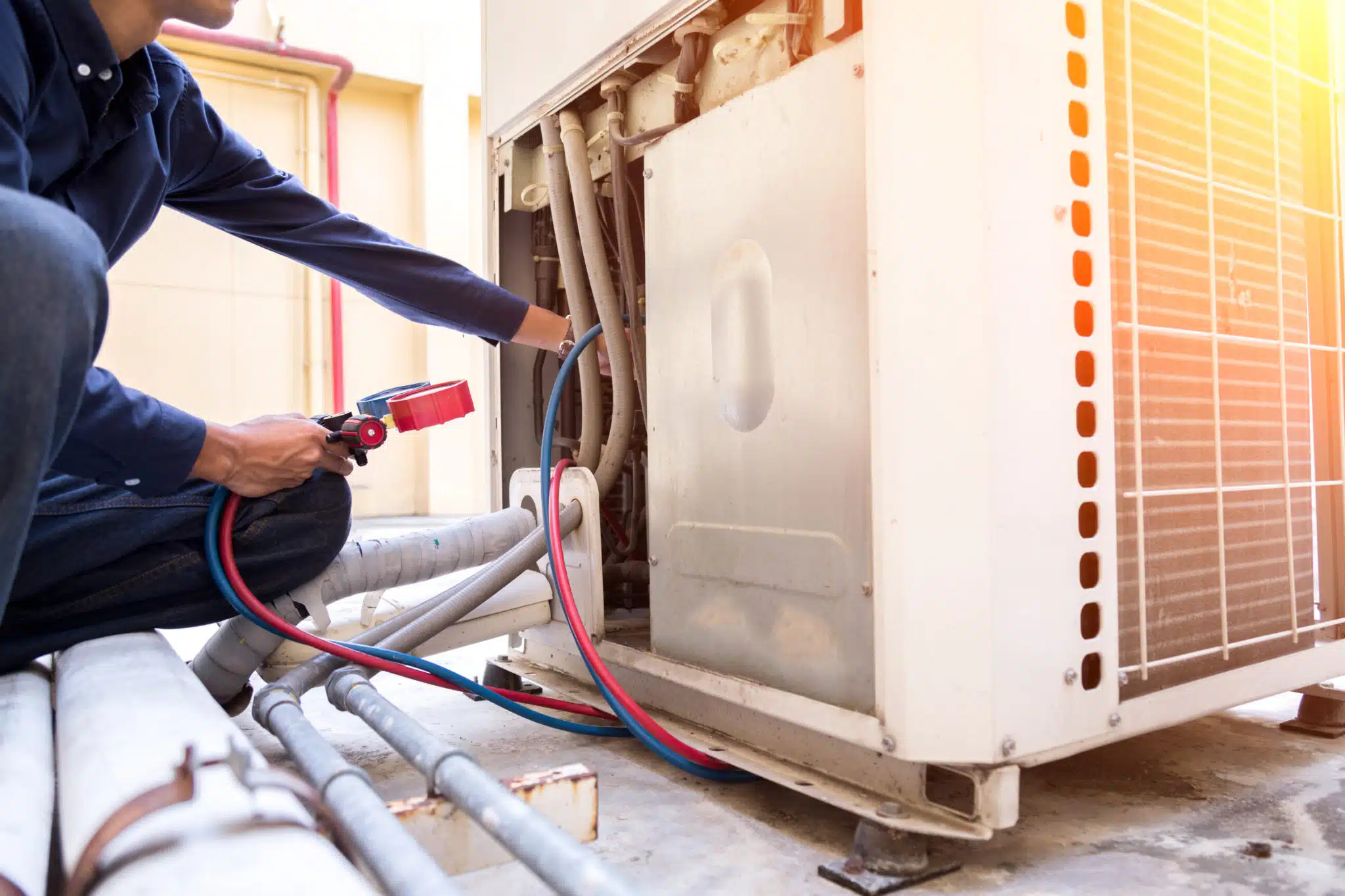 An HVAC technician expertly repairs an industrial air conditioning unit on a rooftop, utilizing gauges and tools with precision. Sunlight gleams off the metal casing, highlighting the network of pipes in the foreground.