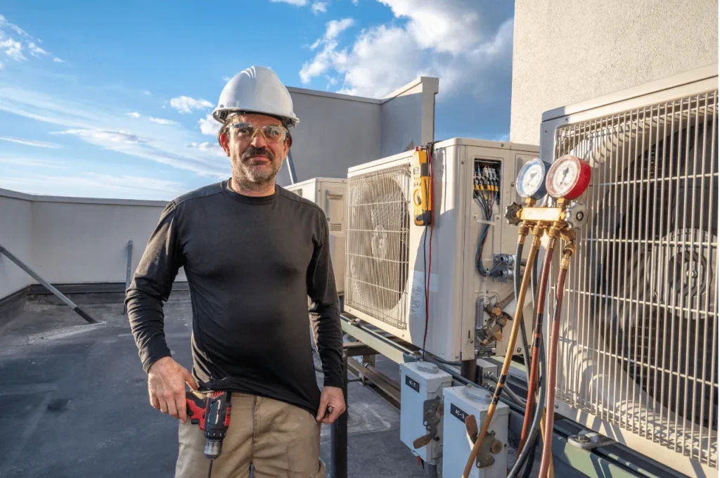 On Hook Agency's website: A worker in a white hard hat and protective eyewear stands confidently on a rooftop beside HVAC units, essential for optimizing profit margins. Equipped with a drill and manifold gauge set connected to the units, he signifies readiness and precision under partly cloudy skies.