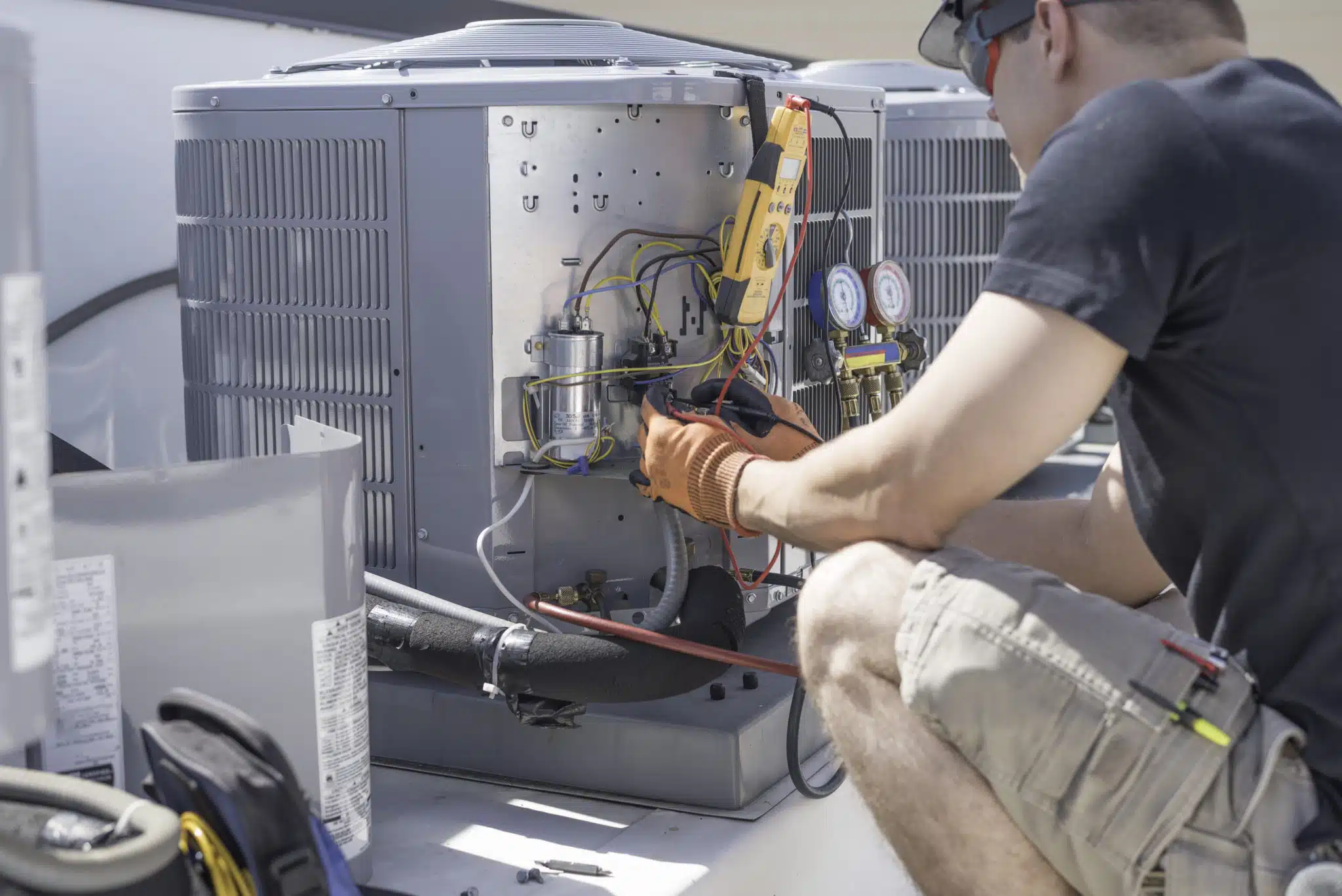 A technician in a black shirt and gloves expertly repairs a rooftop air conditioning unit. Using his HVAC expertise, he employs various tools to inspect the wiring and components, ensuring efficient performance and optimal functionality.