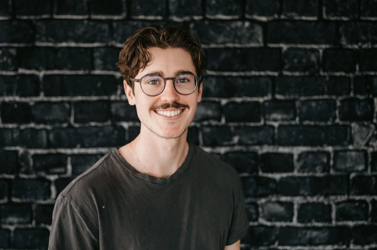 Tim Brown stands confidently in front of a textured dark brick wall, sporting short, wavy hair and a mustache. With glasses and a simple dark t-shirt, his relaxed demeanor adds to the scene's appeal.