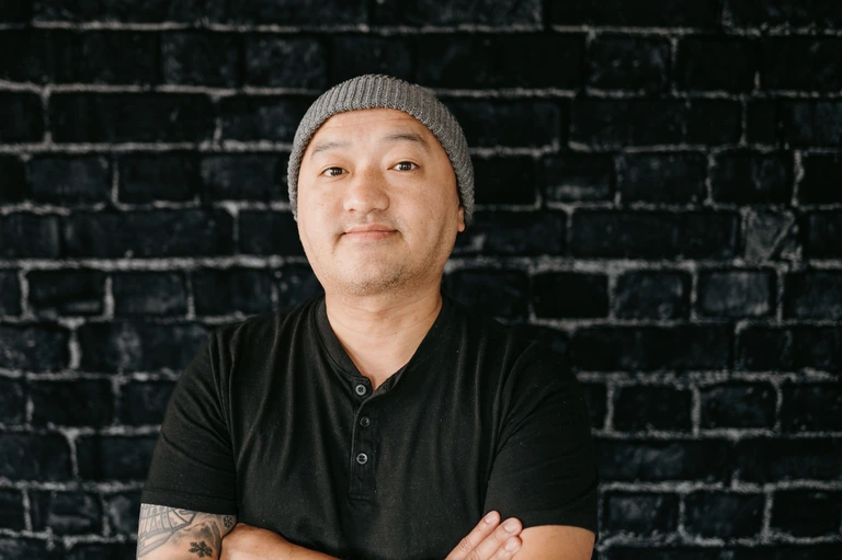 Tim Brown stands confidently before a dark brick wall, wearing a gray beanie and black shirt, arms crossed and smiling at the camera.