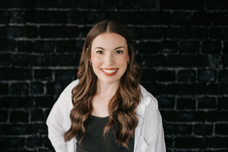 A woman with long brown hair and a bright smile, embodying Tim Brown’s design aesthetic, stands against a dark brick wall in a black top and white open shirt.