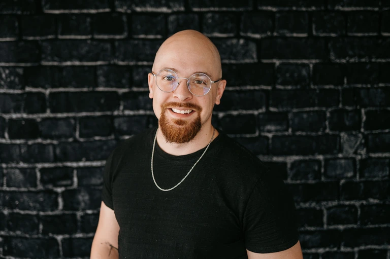 Tim Brown, a man with glasses, a beard, and a bald head, stands confidently in front of a black brick wall. He is dressed in a black T-shirt and sports a silver chain necklace.