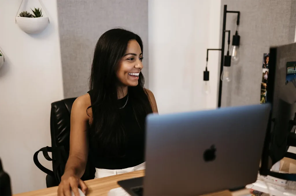 A woman with long dark hair sits at a desk, attentively working on her computer. She wears a black sleeveless top, framed by modern hanging light fixtures and a wall planter in the background.